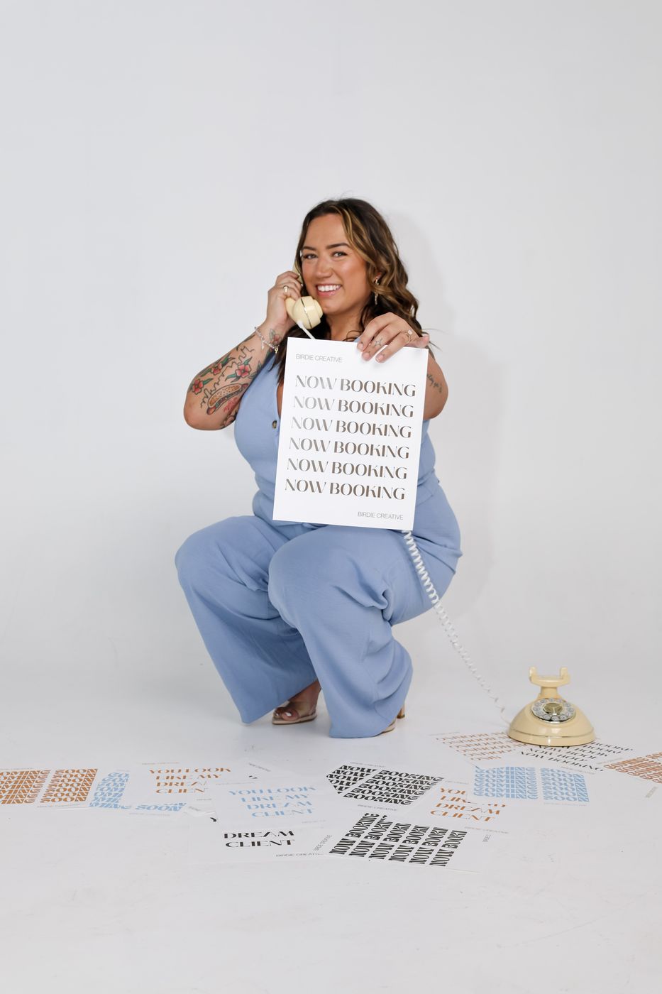 Zoe crouching in a light blue outfit with a Now Booking sign and cream rotary phone