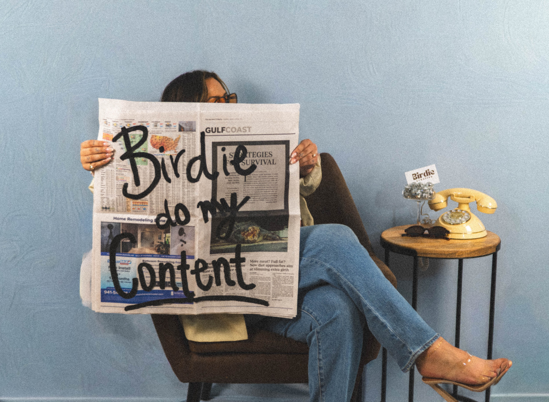 Zoe seated against a light blue wall holding a newspaper reading Birdie do my content, beside a yellow rotary phone