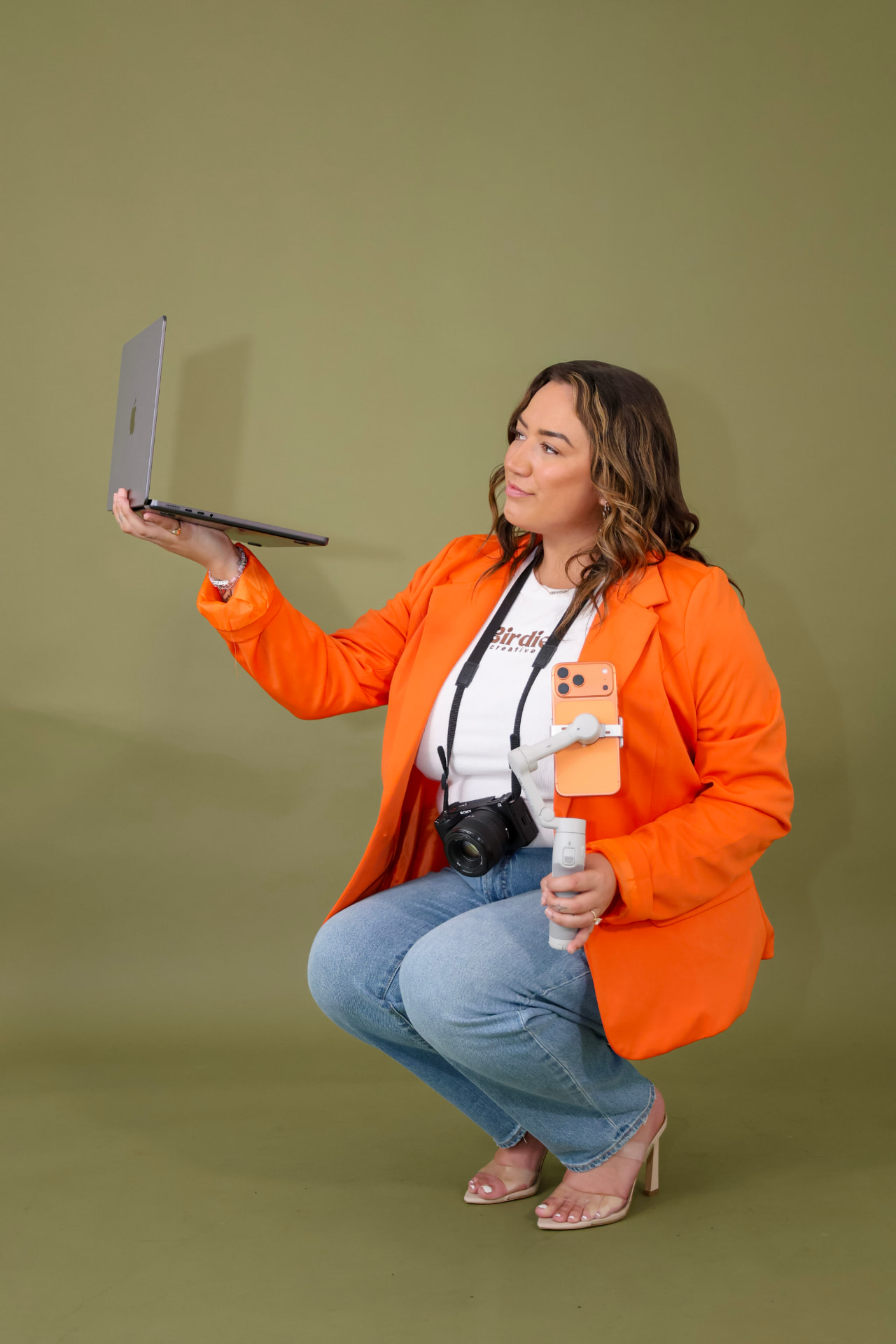 Zoe in an orange blazer holding a laptop, camera and phone stabilizer against an olive backdrop