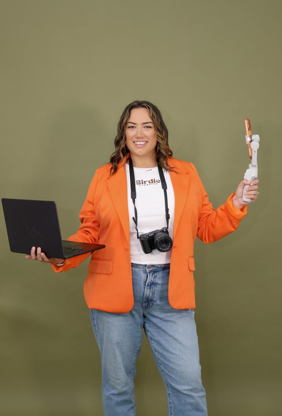 Zoe in an orange blazer with a laptop and gimbal against an olive backdrop