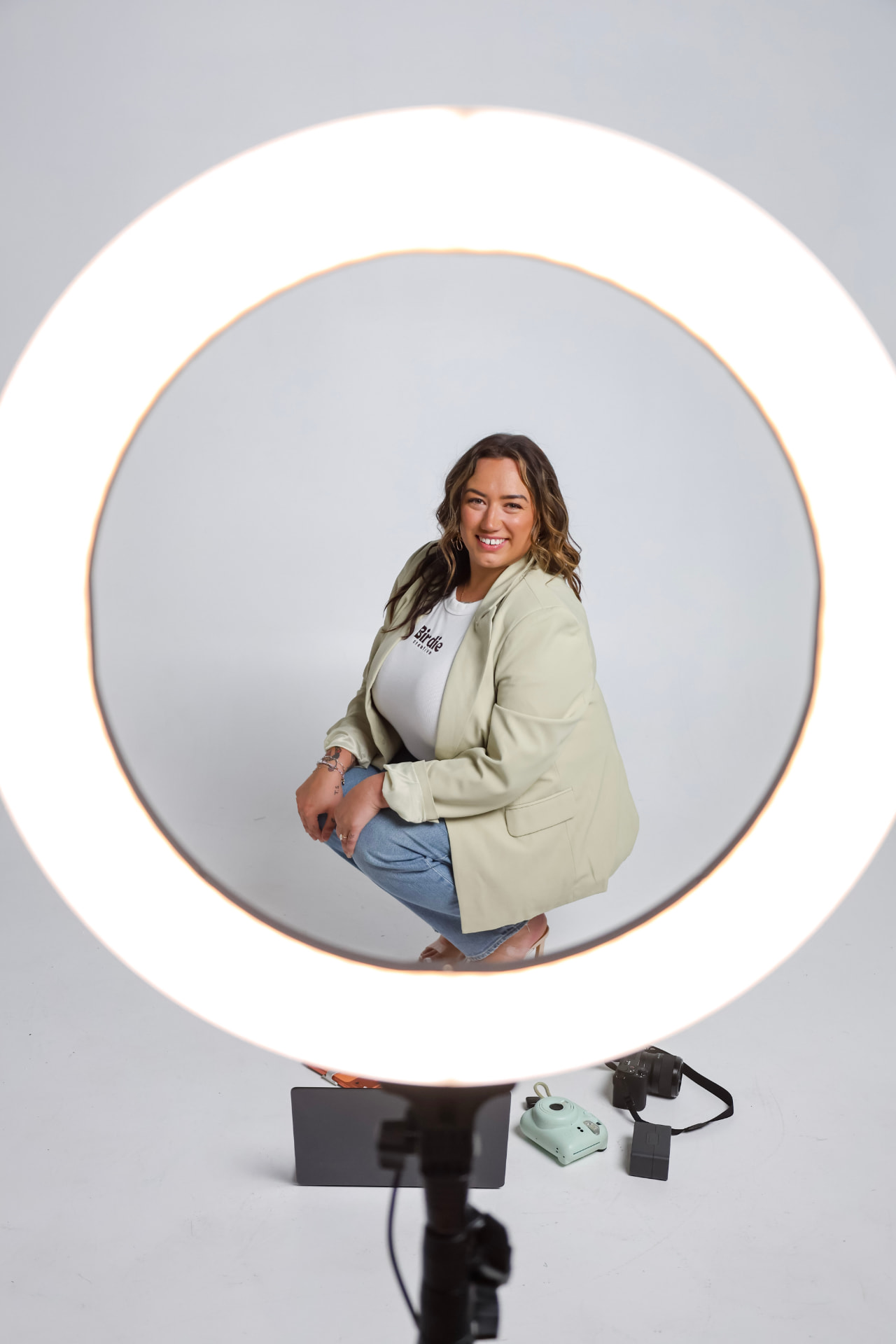 Zoe Kasper seated inside a glowing ring light, smiling in a sage blazer and jeans